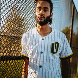 A man near a baseball field fence in a green striped white baseball jersey with green and gold O for Oakland applique on the chest.