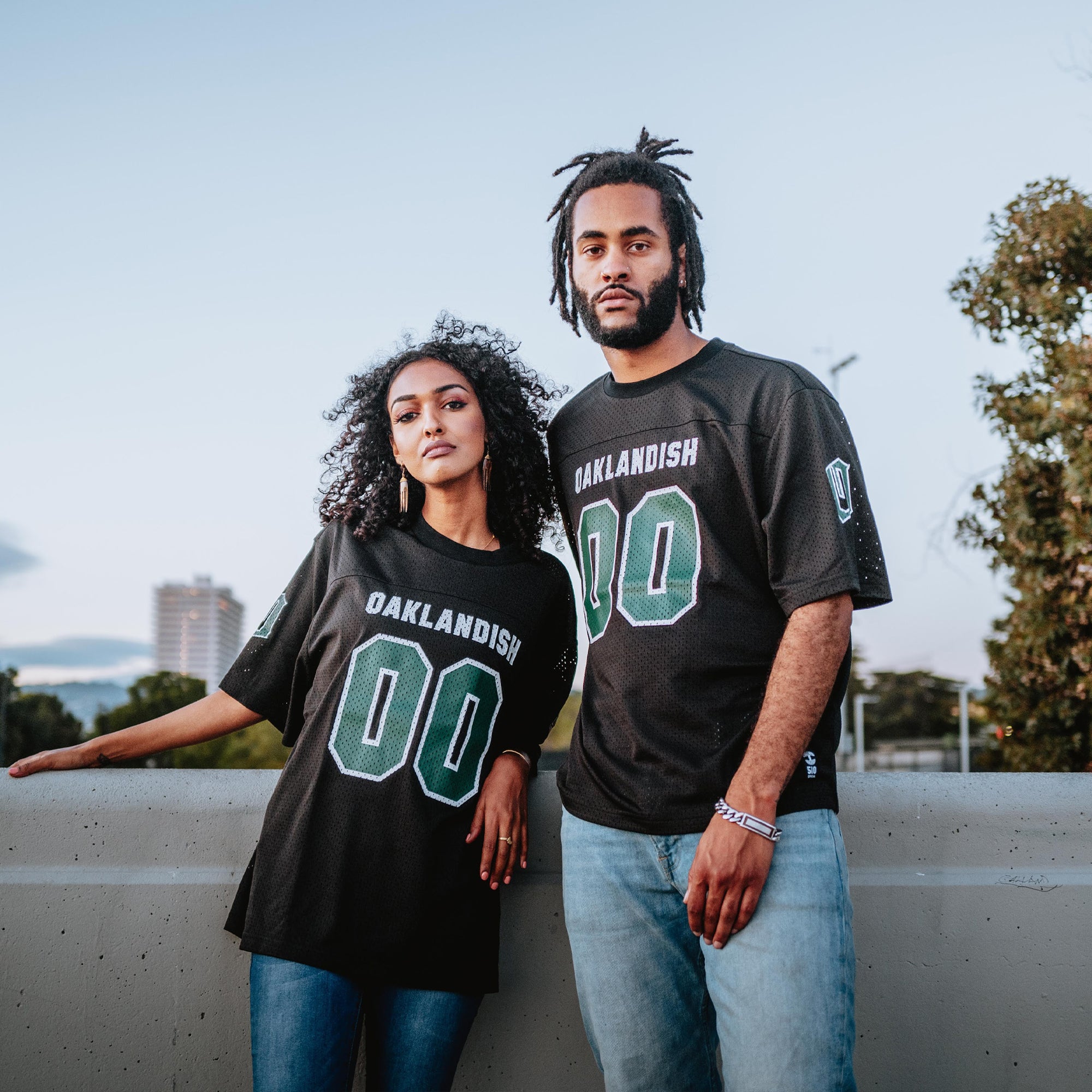 A woman with curly hair and a man stand outdoors in black Other Official Football Home Jerseys, both featuring the number 00. She leans on a concrete ledge while he rests an arm beside her, set against a backdrop of trees and a building under a clear sky.