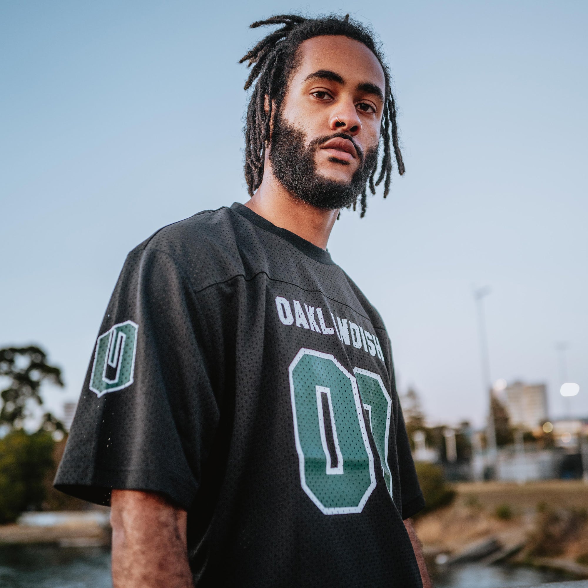 A man with dreadlocks and a beard wears the "Other" brand's Official Football Home Jersey. This black mesh jersey features "OAKLANDISH" and number "01" in white and green. He's outdoors against a blurred backdrop of trees, buildings, and clear sky.