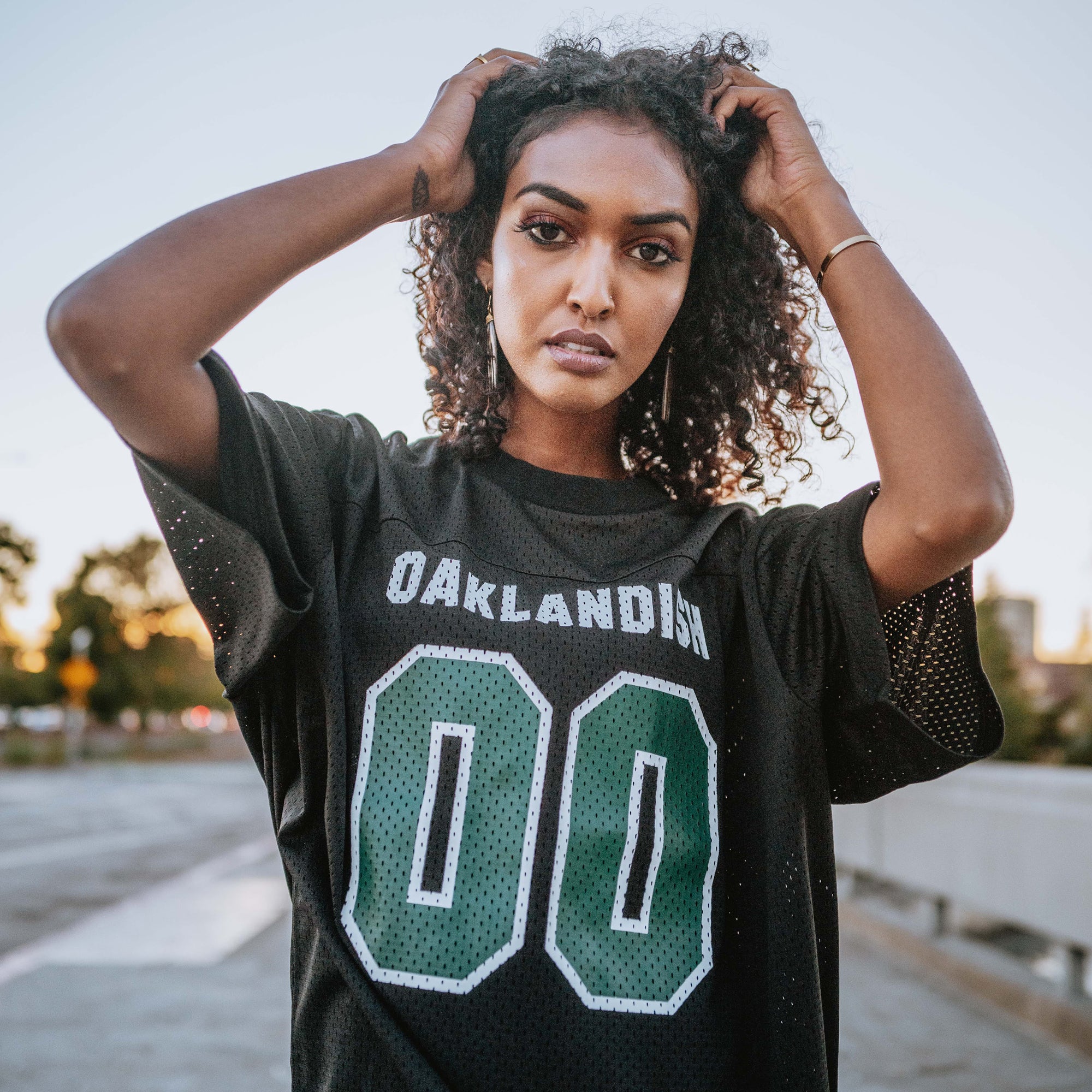 Woman outside in a black mesh football jersey with 00 green numbers and large white Oaklandish wordmark.