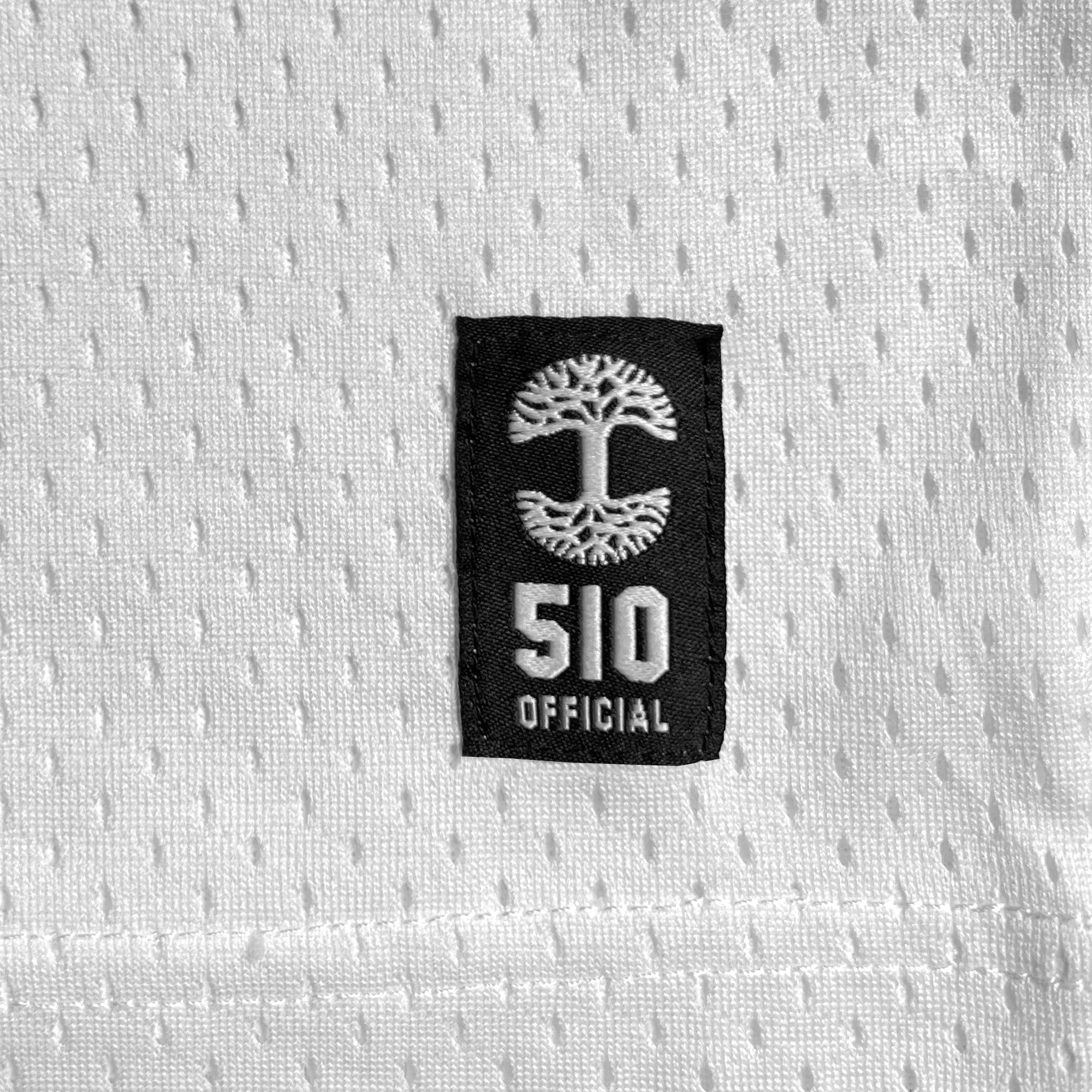 A close-up of the Official Football Away Jersey shows white textured mesh fabric with small perforations, featuring a black patch with an Oaklandish-style white tree emblem and bold "510 OFFICIAL" text. Visible stitching lines the bottom edge. Produced by brand Other.