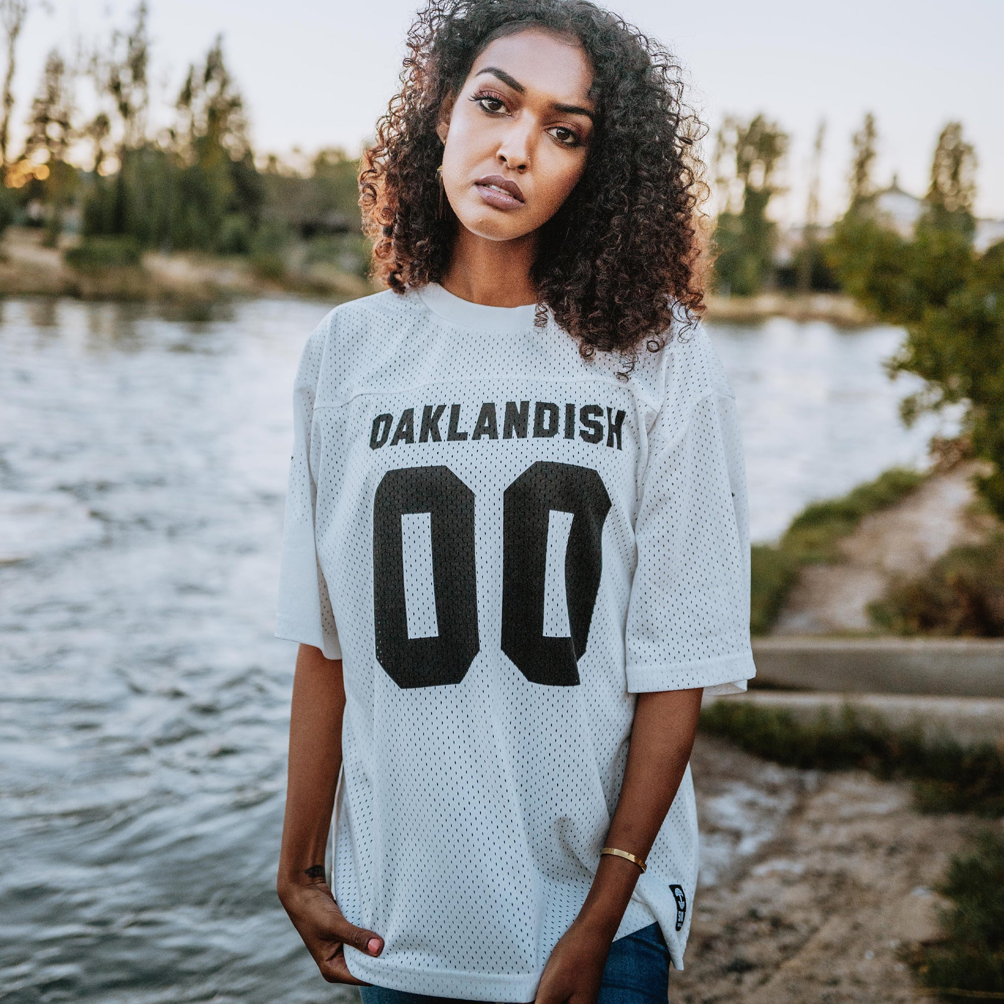A woman stands by a riverbank wearing the lightweight Official Football Away Jersey by Other, marked "OAKLANDISH 00" in bold black. Her curly hair frames a neutral expression while trees and calm water create a serene backdrop under an overcast sky.