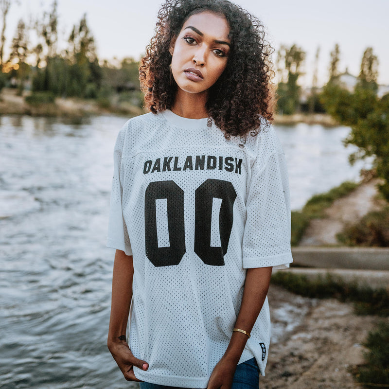 A woman standing outside in a white mesh football jersey with 00 black numbers and a large black Oaklandish wordmark.