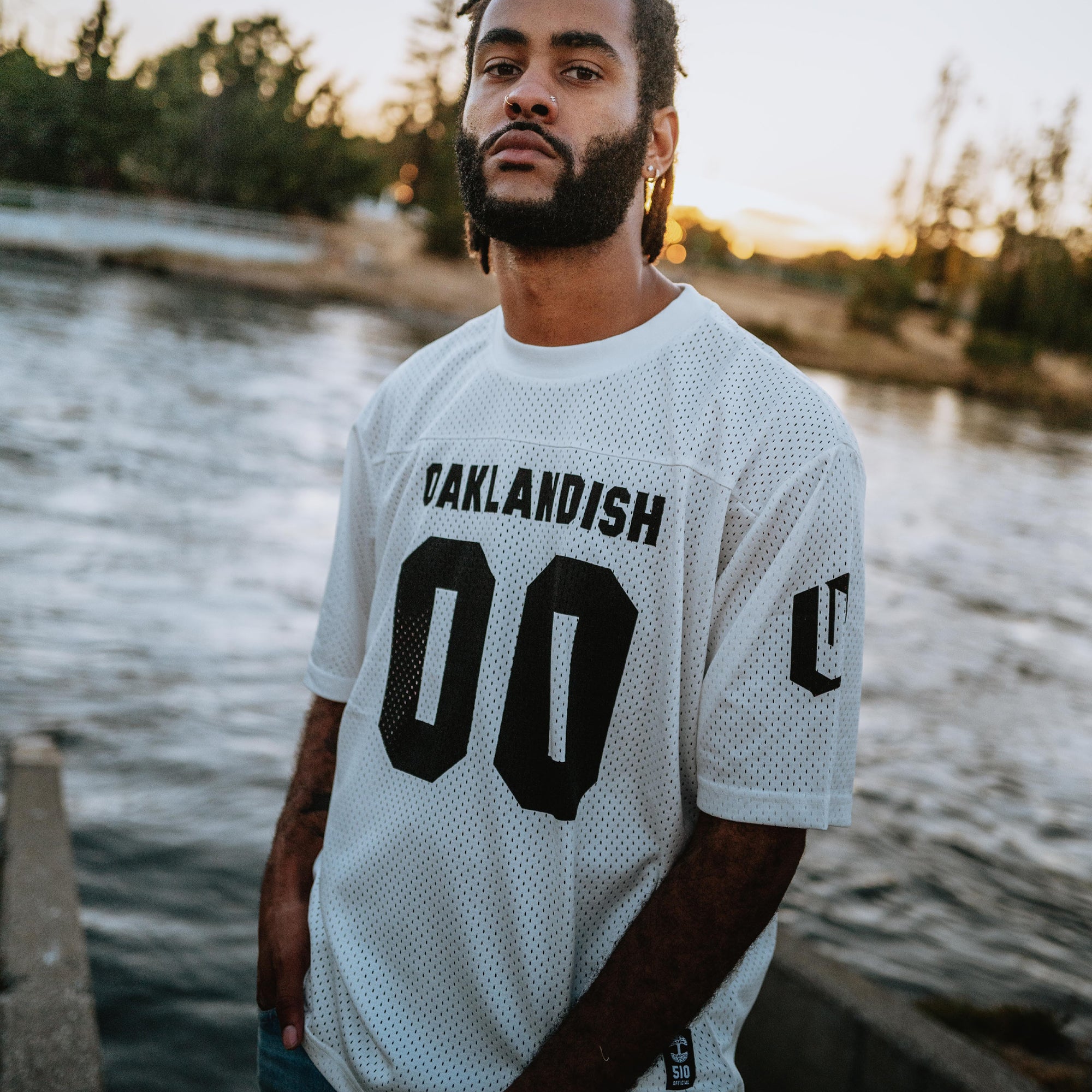 A man standing outside in a white mesh football jersey with 00 black numbers and a large black Oaklandish wordmark.