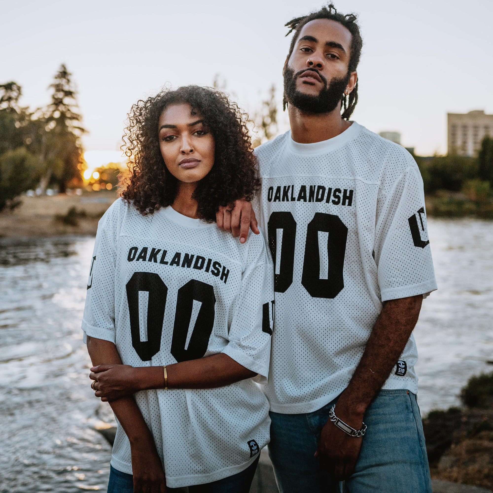 A man with dreadlocks and a woman with curly hair sport serious expressions by a river at sunset, dressed in the "Other Official Football Away Jersey" featuring "OAKLANDISH 00" in black. Clad in jeans, they stand amidst trees and a blurred cityscape.