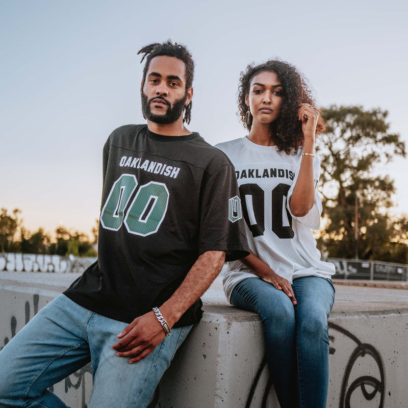 A woman and man standing outside in mesh Oaklandish football jerseys. 