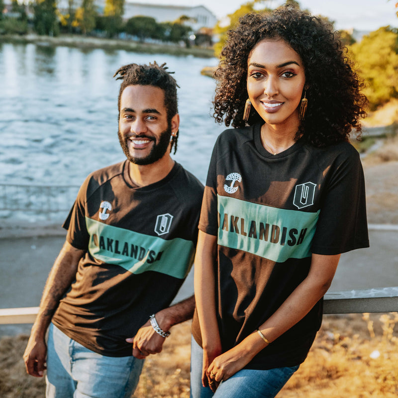 Man and woman wearing black and green striped Oaklandish soccer jerseys.