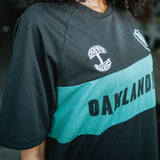 Close-up of OAKLANDISH wordmark, tree logo, and O in the green stripe on a woman’s chest.