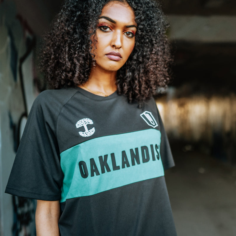Woman wearing black soccer jersey with green stripe with black OAKLANDISH wordmark, white Oaklandish tree logo, and O appliques.
