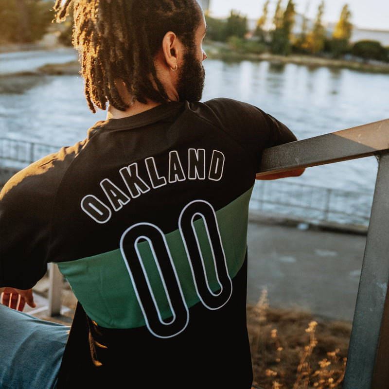Man outside, back to the camera in a black soccer jersey with a green stripe and OAKLAND 00 applique.