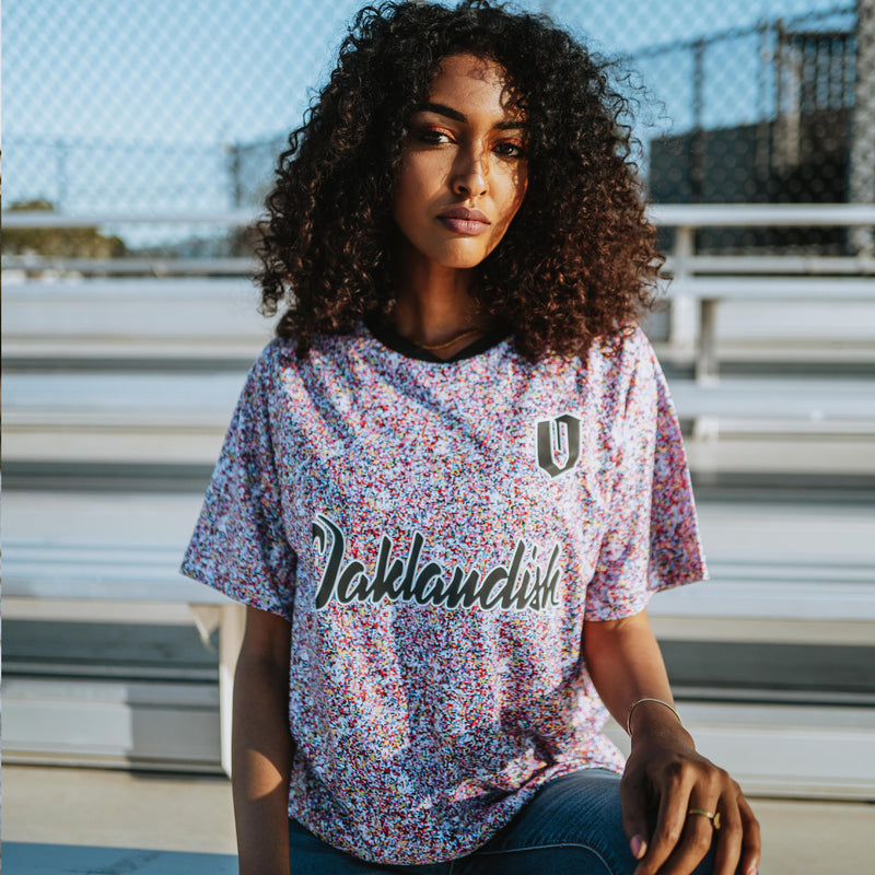 Women wearing multi-colored speckled soccer shirt with cursive Oaklandish wordmark and black O for Oakland applique.