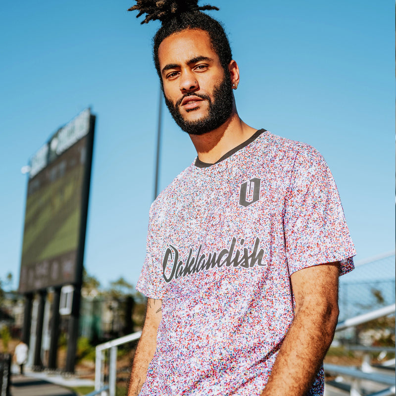 Man wearing multi-colored speckled soccer shirt with cursive Oaklandish wordmark and black O for Oakland applique.