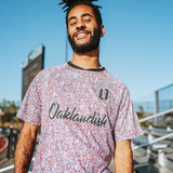 Man wearing a multi-colored speckled soccer jersey with cursive Oaklandish wordmark and black O for Oakland applique in bleechers.