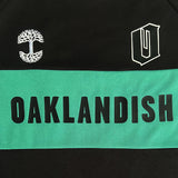 Close-up of OAKLANDISH wordmark, tree logo, and O in the green stripe on a black soccer jersey.