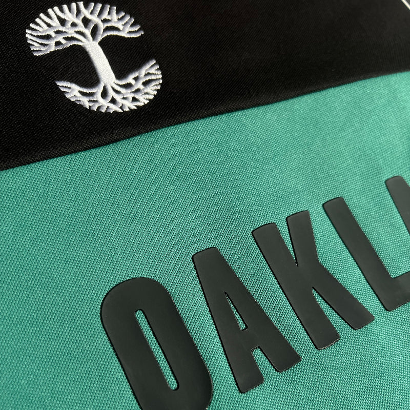 Close-up of half of OAKLAND wordmark and white embroidered tree logo on a black and green soccer jersey.