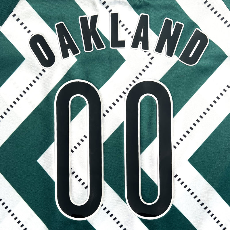 Close-up of black OAKLAND 00 wordmark on the back side of a soccer jersey with a green and white zig-zag pattern.