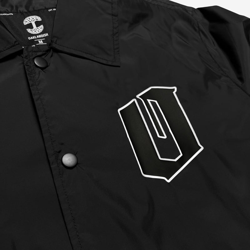 Close up of black and white Oaklandish O logo on the chest of a black nylon coaches jacket. 
