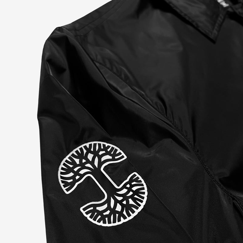Close up of black and white Oaklandish tree logo on the sleeve of a black nylon coaches jacket. 