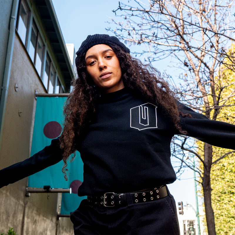 A woman outdoors in Oakland in a black crewneck sweatshirt with an O for Oakland applique patch on the chest.