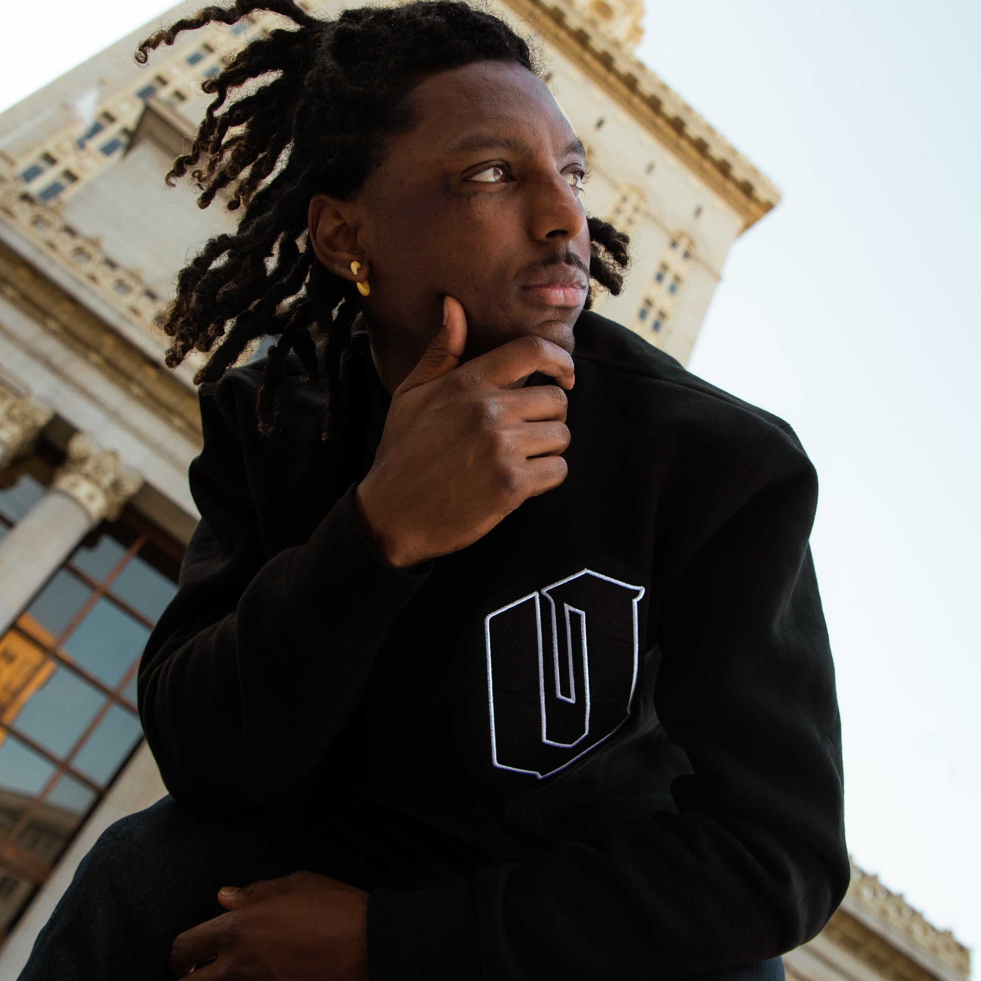 A man outdoors in Oakland in a black crewneck sweatshirt with an O for Oakland applique patch on the chest.