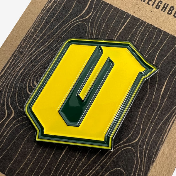 Close up of a green and yellow enamel lapel pin with Official O for Oakland on brown paper Oaklandish retail packaging.