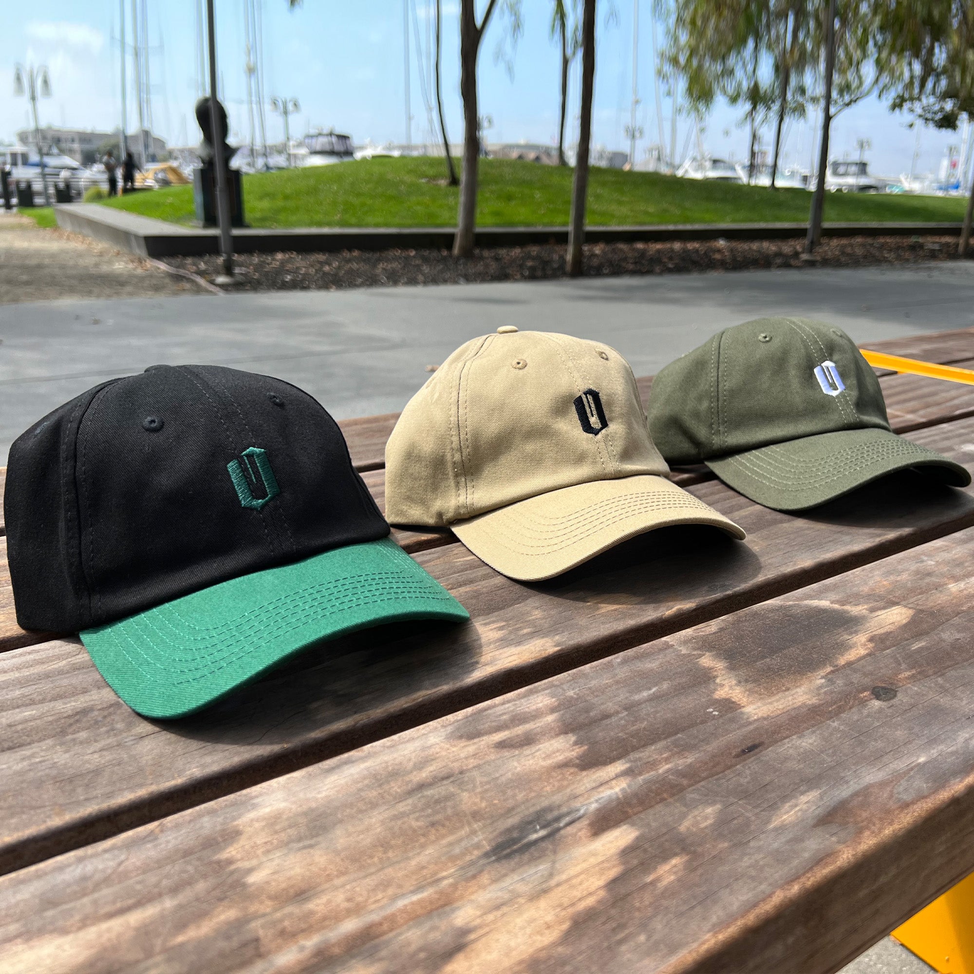 Three baseball caps are placed side by side on a wooden bench outdoors, with a marina in the background. The caps are black with a green brim, beige, and olive green. Among them is an olive green dad hat from Oaklandish, featuring an embroidered "O" and named the Official Micro Logo Dad Hat. Tree-lined green spaces and parked boats add to the serene setting.