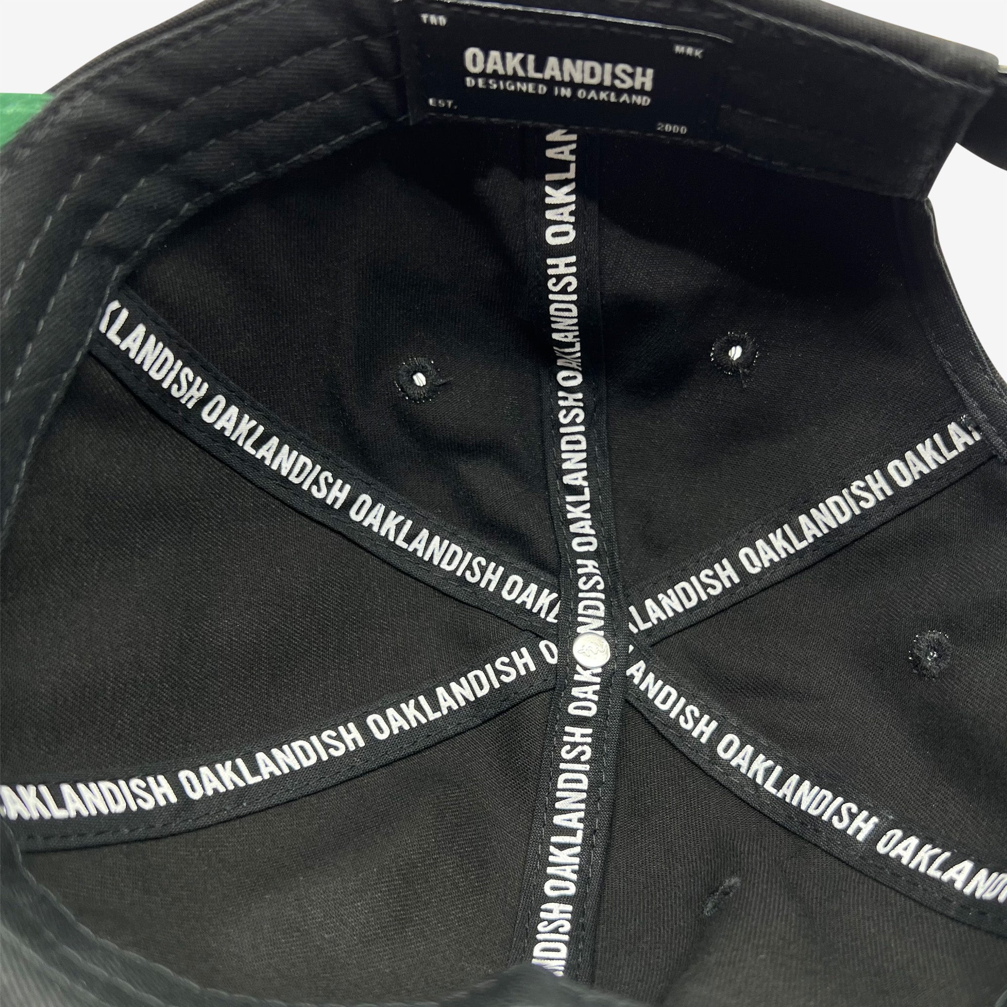 The interior view of the Official Micro Logo Dad Hat in black showcases the brand "Oaklandish" printed repeatedly along the inner seams. A label on the inside reads "Oaklandish Designed in Oakland Est. 2000." The cap features an embroidered "O" and multiple triangular sections converging at the top button.