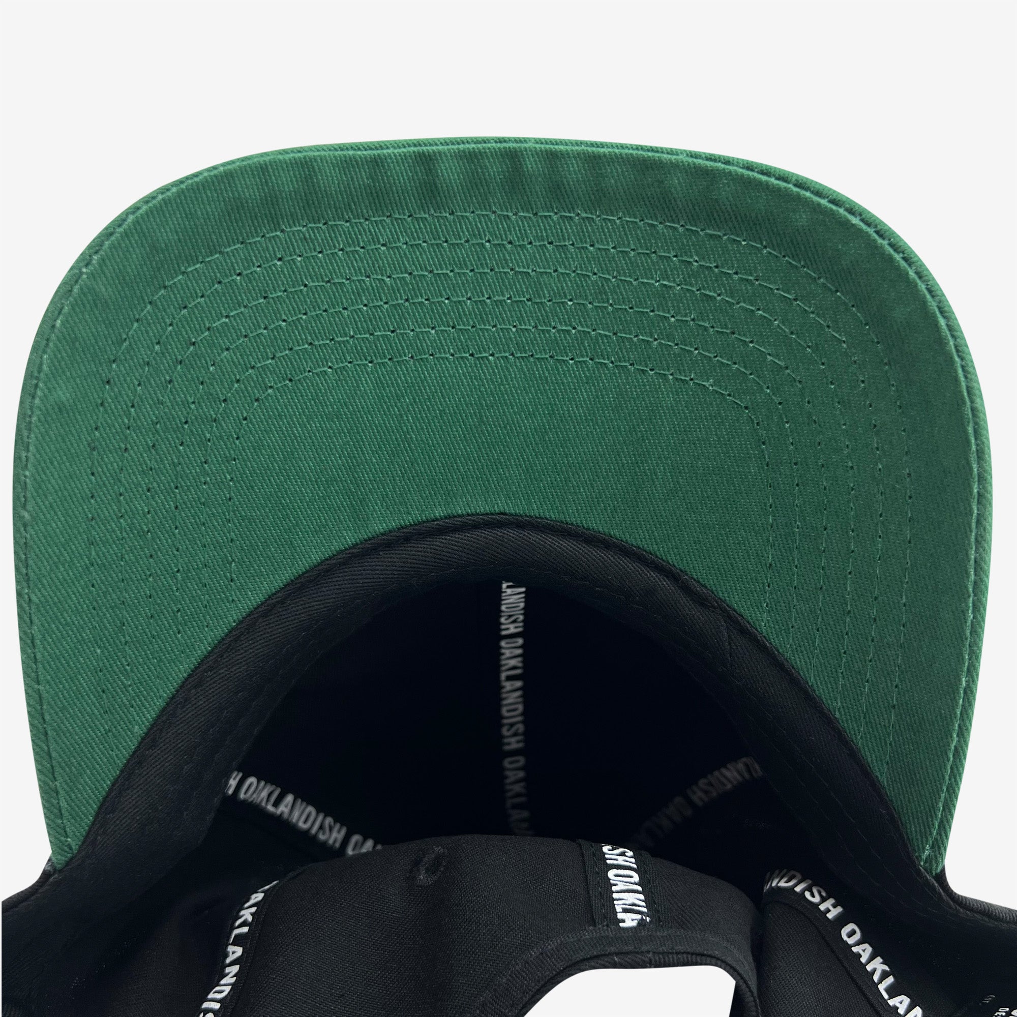 A close-up view of the underside of the Official Micro Logo Dad Hat's green brim. The inner band is black, adorned with "OAKLANDISH" repeatedly printed in white. From this bottom-up perspective, you can also notice an embroidered "O," adding a unique touch to the Oaklandish design.