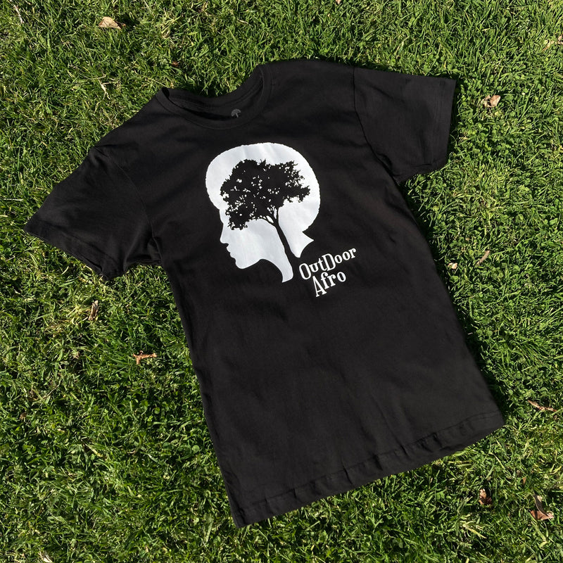 Black t-shirt with white OutDoor Afro logo and wordmark lying outdoors on grass.