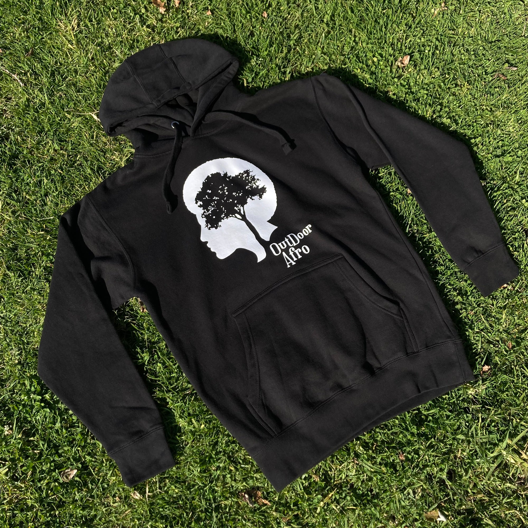 The Outdoor Afro Hoodie by Partner is a premium heavyweight fleece pullover that showcases a white silhouette of a person's profile on the chest. Inside the silhouette, an image of a tree represents the person's hair, and below this design, "Outdoor Afro" is printed. The hoodie is displayed on green grass.