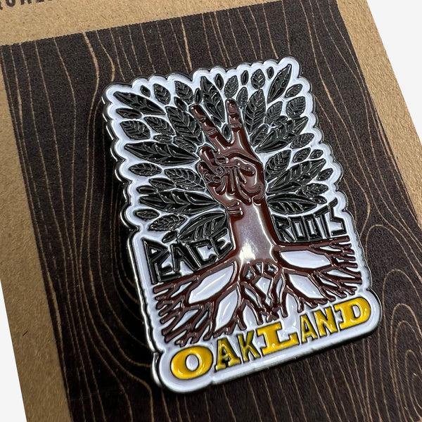 Close-up with enamel pin with Oaklandish wordmark, brown roots, peace sign hand, green leaves, and Peace and Roots.