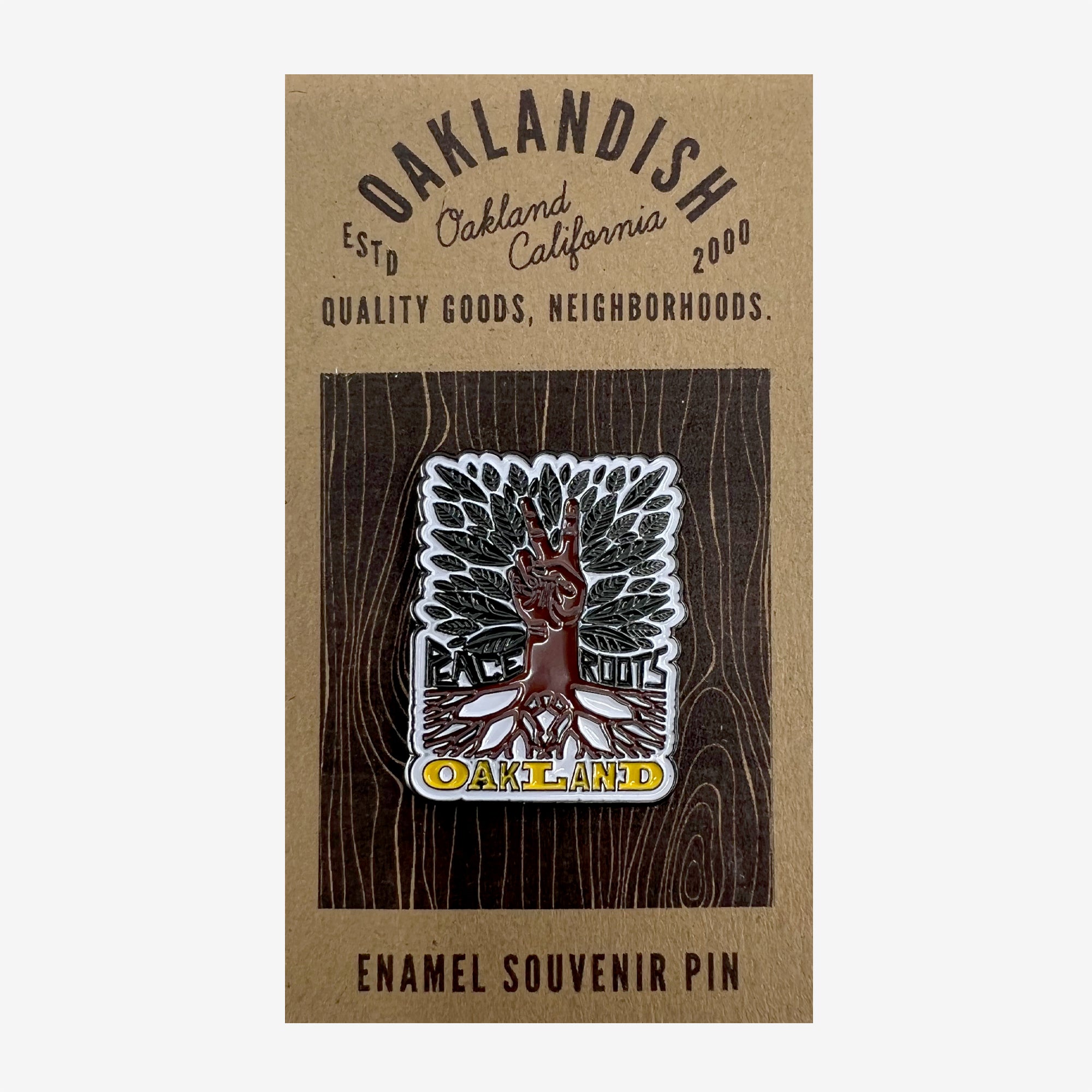 The "Enamel Pin - Peace and Roots" from Oaklandish captures the essence of Oakland with a rectangular design. It features a tree with roots encircling a raised hand making a peace sign, and "OAKLAND" prominently displayed below. This accessory is mounted on a brown card that includes the "OAKLANDISH" branding and additional descriptive text.