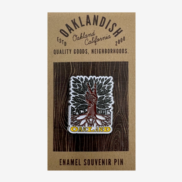 Enamel pin with yellow Oaklandish wordmark, brown roots, peace sign hand, green leaves, and Peace and Roots on retail packaging.