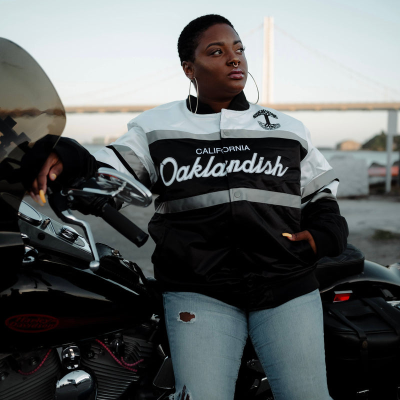 Woman on a motorbike in a striped, collared satin jacket with a California Oaklandish wordmark and Oaklandish tree logo.