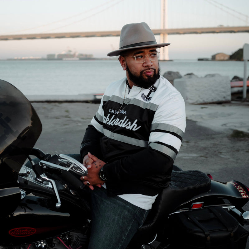 A man sitting on a motorbike in a striped satin jacket with a California Oaklandish wordmark and Oaklandish tree logo.
