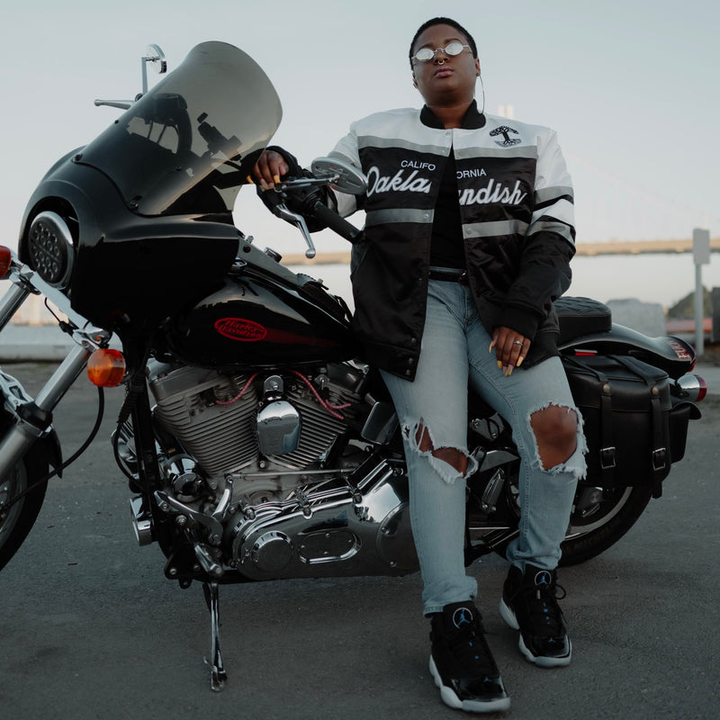 A woman sitting on a motorbike in a striped satin jacket with a California Oaklandish wordmark and Oaklandish tree logo.
