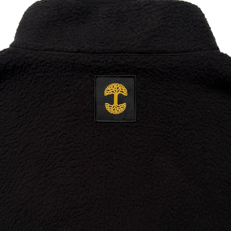 Close-up of a yellow Oaklandish tree logo patch under the collar of a black polar fleece pullover.