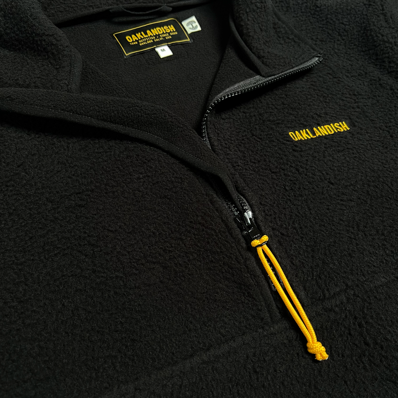 Close-up of yellow embroidered Oaklandish word mark, ½ zip with yellow pull and inside neck logo, on a black polar fleece pullover.