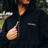 Close-up of yellow embroidered Oaklandish word mark and ½ zip with yellow zip pull on a woman outdoors in a black polar fleece pullover.