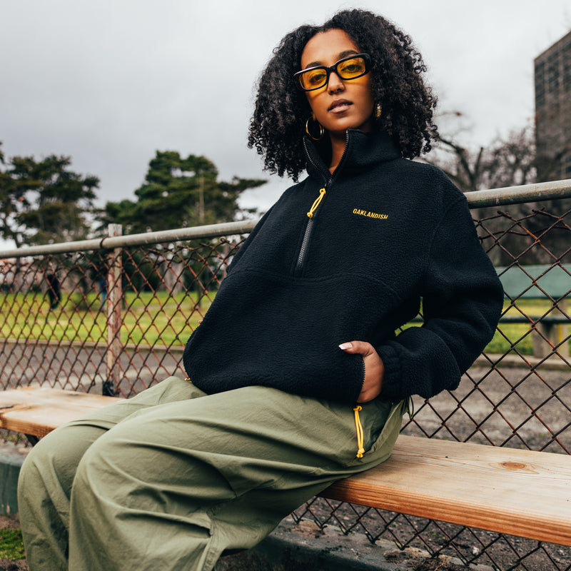 A woman sitting on outdoor bleachers, hands in pockets,  in a black polar fleece pullover with 1/2 zip, yellow zip pulls, and a yellow Oaklandish wordmark.
