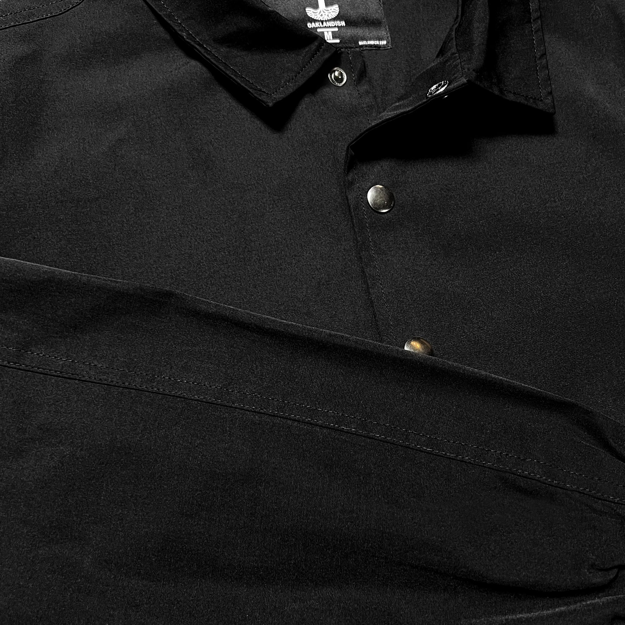 Close-up of the Oaklandish Premium Coaches Jacket in black. The image showcases the collar and part of the upper section, focusing on the metallic buttons. The matte fabric appears smooth. The top button is fastened, while the second button remains unbuttoned. The sleeve is partially visible, hinting at its men's sizing.