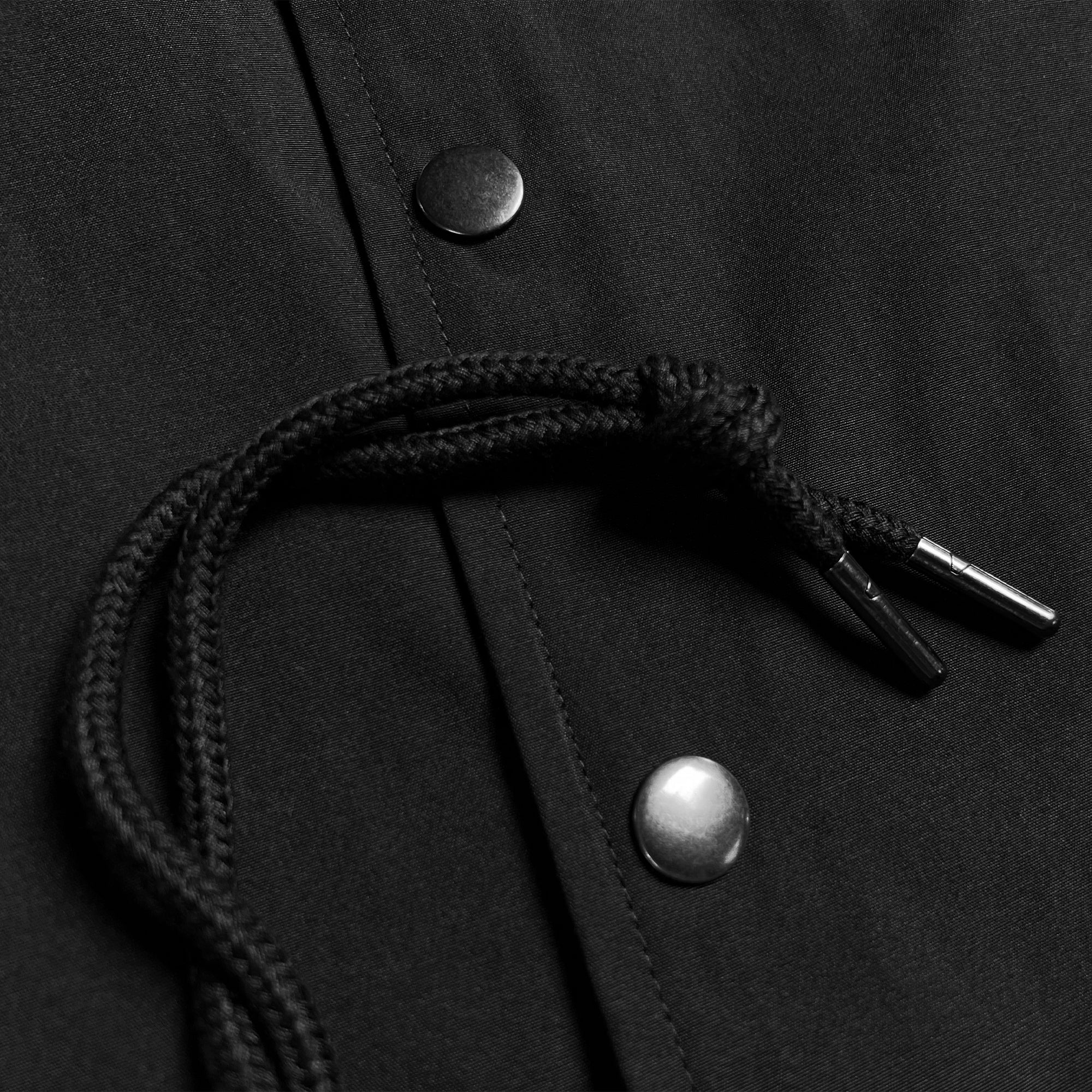 A close-up image of Oaklandish's Premium Coaches Jacket in black, highlighting metallic snap buttons and a braided black drawstring with metal aglets at the ends. The jacket's material appears smooth, slightly reflective, and water-resistant. The focus is on the fine details of the buttons and drawstring.