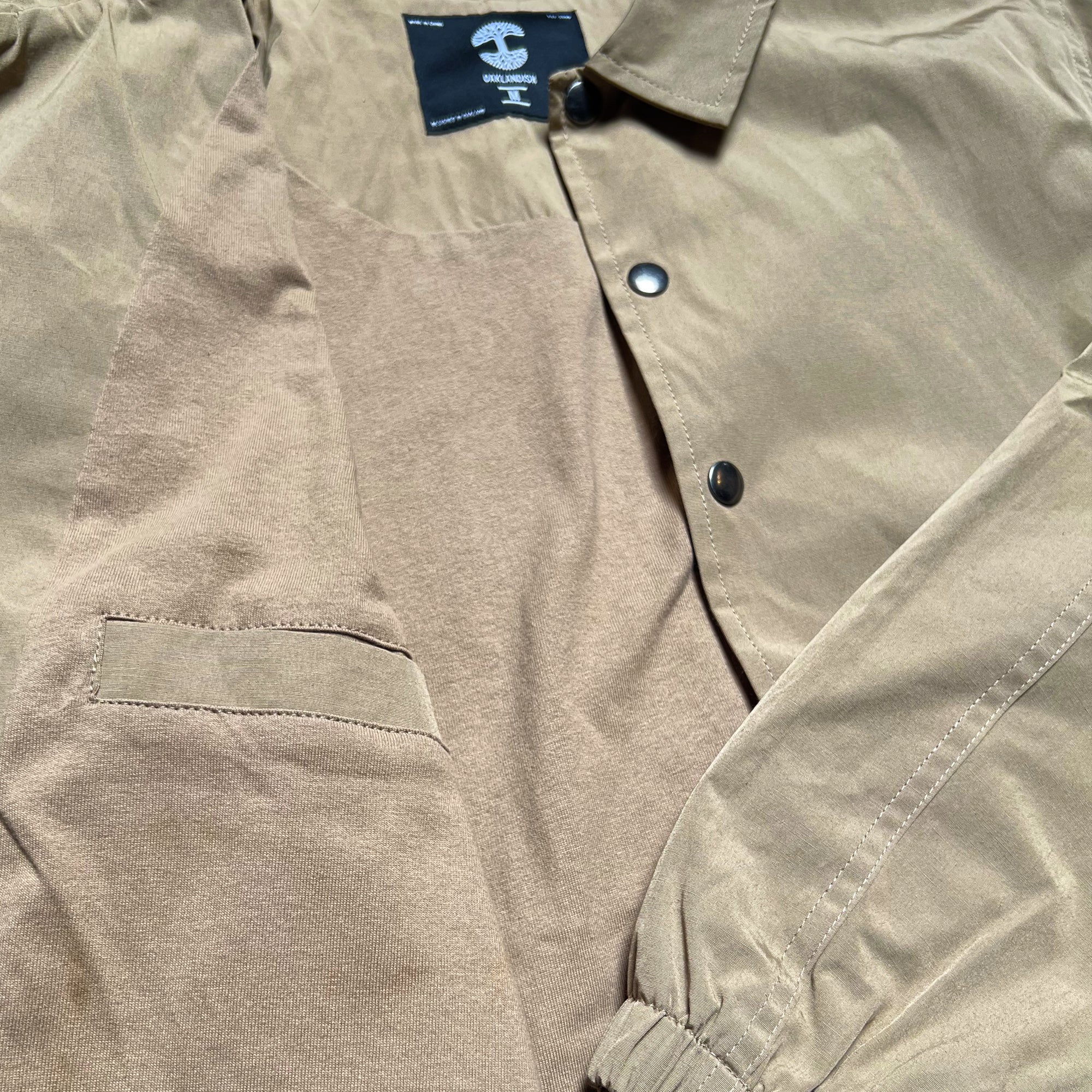 A tan, lightweight windbreaker jacket is shown lying flat. This Oaklandish Premium Coaches Jacket features a black snap closure, elastic cuffs, and a pocket on the left inner side. A black label at the collar displays brand information and men's sizing. The inner lining contrasts slightly with the outer fabric's texture.