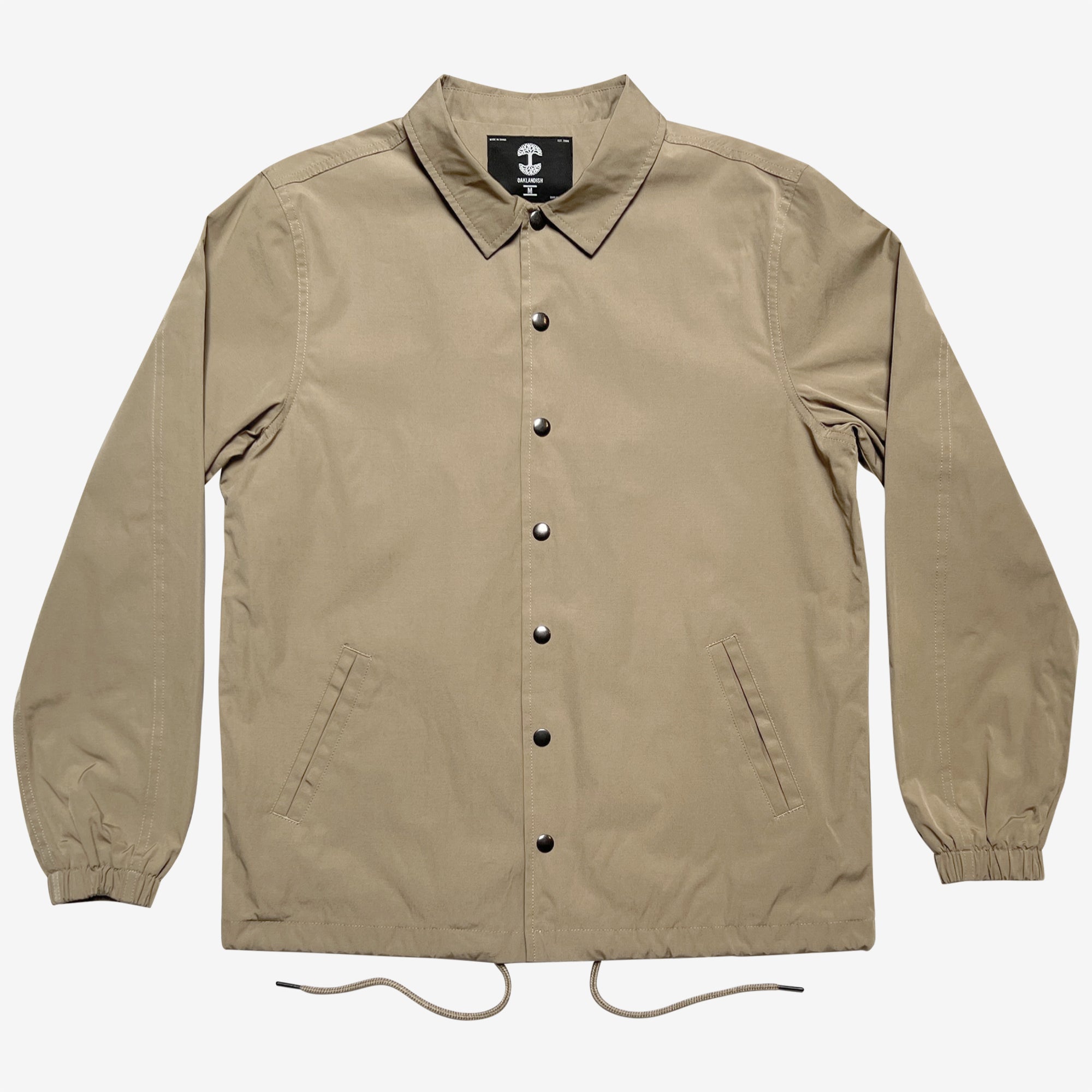 The Oaklandish Premium Coaches Jacket is a beige, long-sleeve jacket featuring a collar and elastic cuffs. This water-resistant jacket includes a drawstring hem, diagonal front pockets, and black snap buttons down the front. The interior label displays a white anchor symbol on a black background. The men's sizing jacket is shown laid flat against a white backdrop.