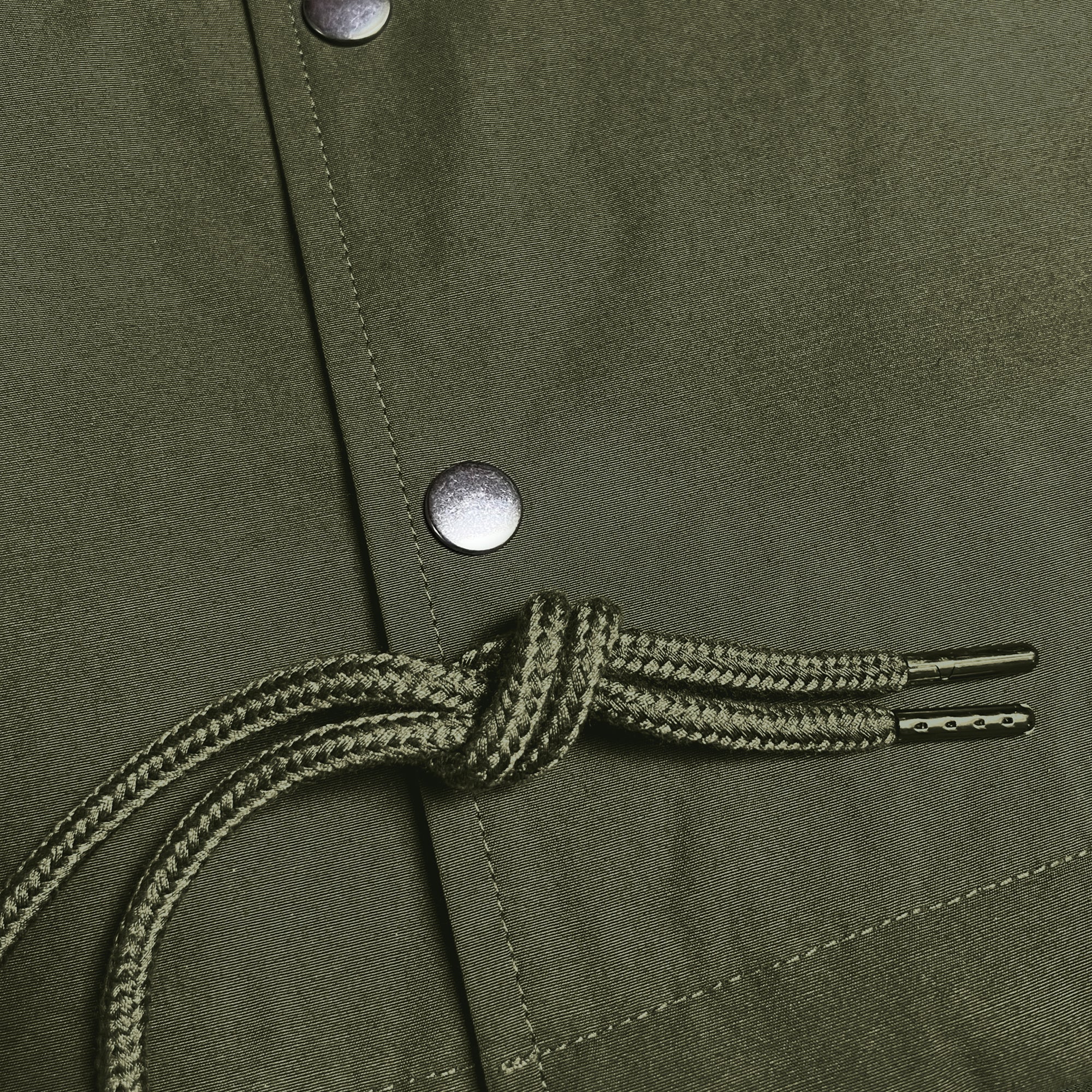 Close-up of a khaki green fabric with a drawstring featuring two black aglets tied in a knot. The fabric is part of the Premium Coaches Jacket by Oaklandish, showcasing visible stitching details and silver snap buttons. The texture and weave of this water-resistant outerwear material are clearly visible in the image.