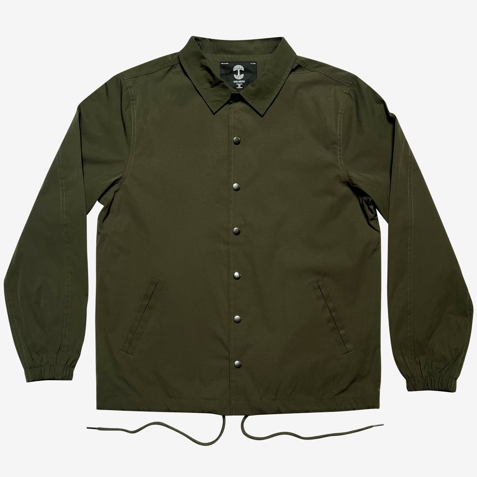 This image showcases the Premium Coaches Jacket by Oaklandish in olive green, featuring long sleeves and a pointed collar. The front displays a row of metallic snap buttons and two diagonal pockets. The elastic cuffs and dual drawstrings at the bottom hem add functionality to this water-resistant outerwear, which exudes a sporty and casual style.