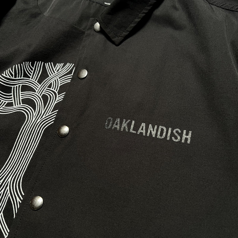 Detailed close-up of large white Oaklandish wordmark on the left chest wearside of black snap close coaches jacket.