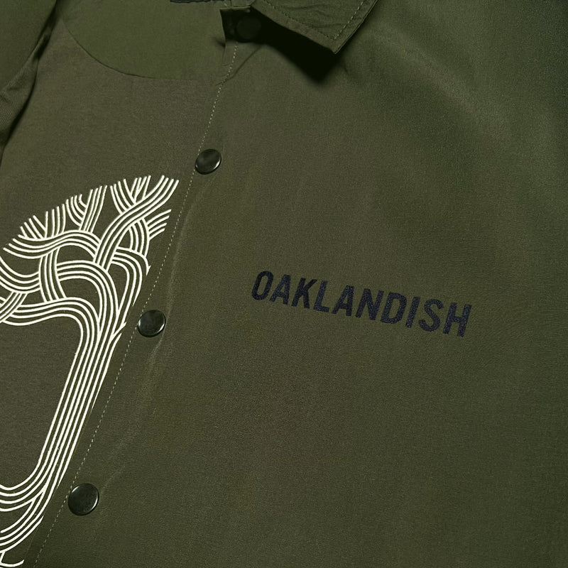 Detailed close-up of black Oaklandish wordmark on the left chest wearside of olive green snap close coaches jacket.