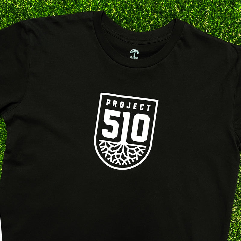 Top view of a black t-shirt with white project 510 Oakland SC Roots logo on grass.