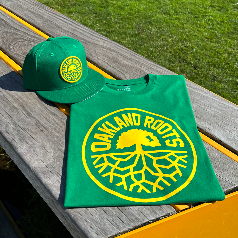 Image of kelly green t-shirt with Oakland Roots SC crest in yellow and Kelly green New Era snapback cap on outdoor picnic table.